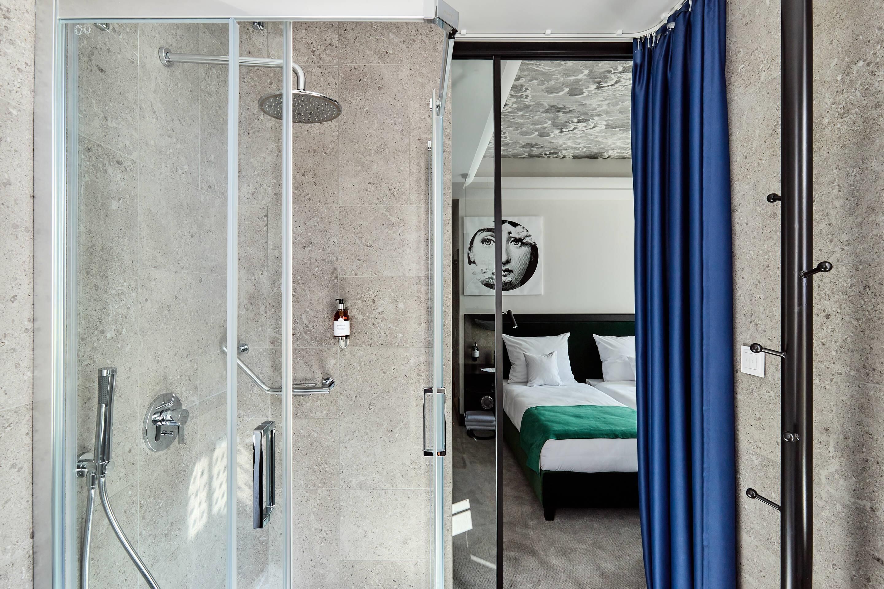 A hotel room with a glass shower enclosure, a bed with white bedding, and a green throw blanket.