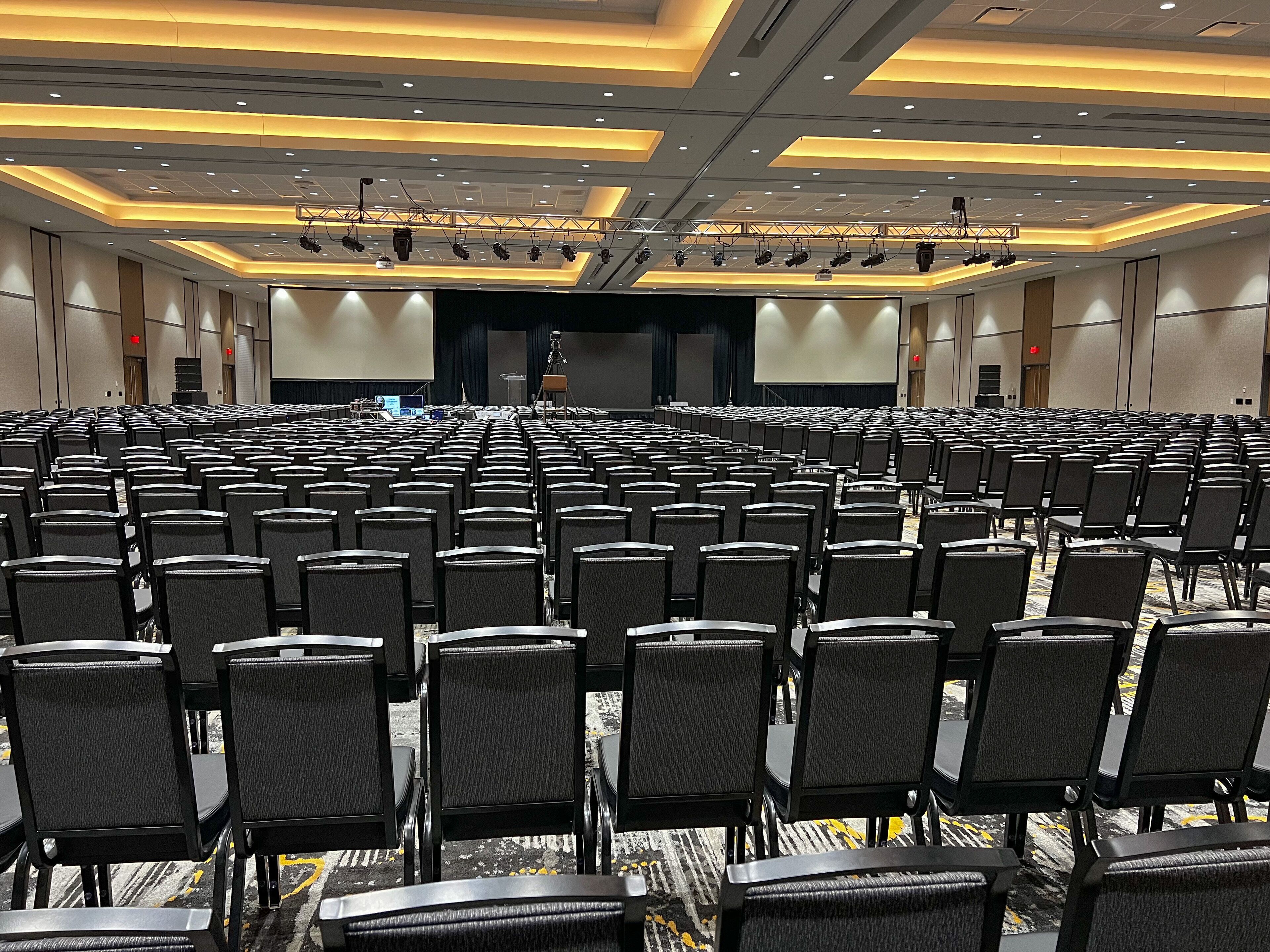 A large conference room with rows of black chairs, a stage with a large screen, and ceiling-mounted lighting.