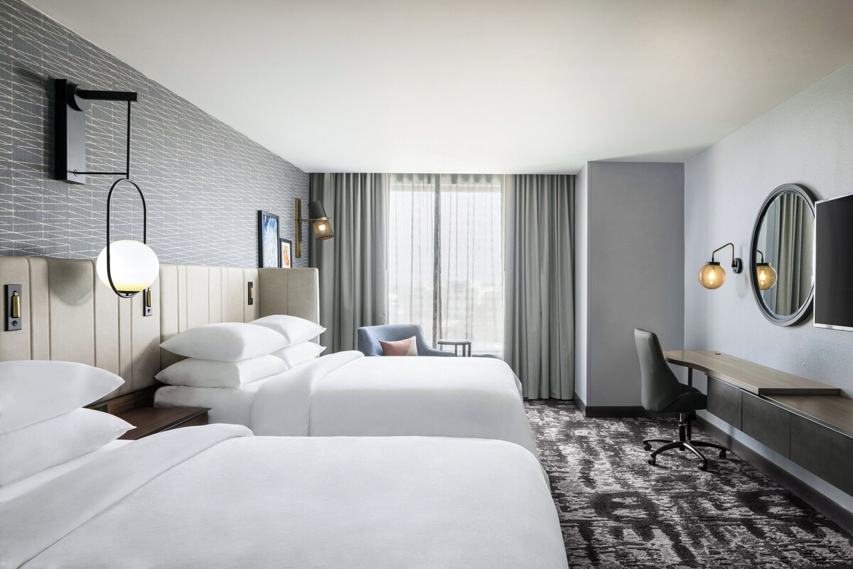 Quarto de hotel com duas camas, uma escrivaninha, uma cadeira, um espelho grande e uma janela com cortinas.