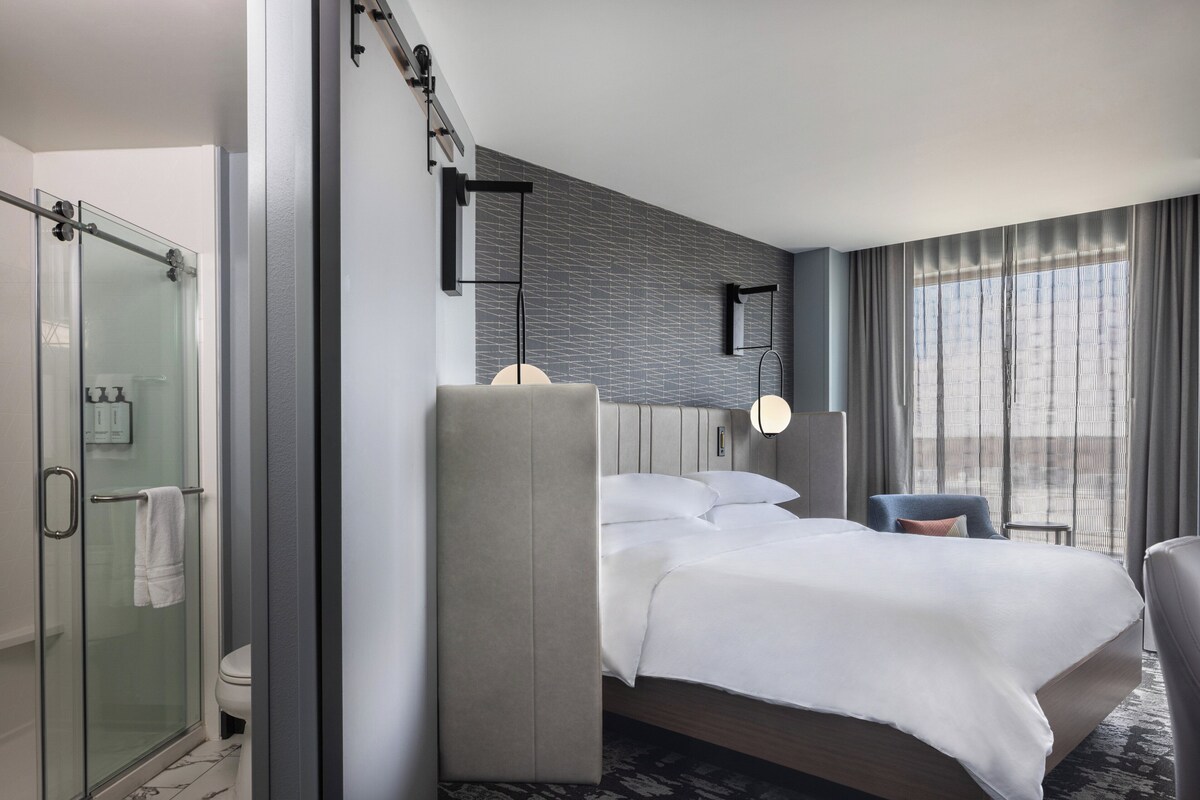 Quarto de hotel moderno com uma cama grande, box de vidro para o chuveiro e vista da cidade pela janela.
