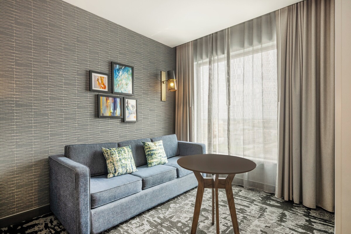 Uma sala de estar moderna com um sofá cinzento, uma mesa de madeira redonda e uma grande janela com cortinas.