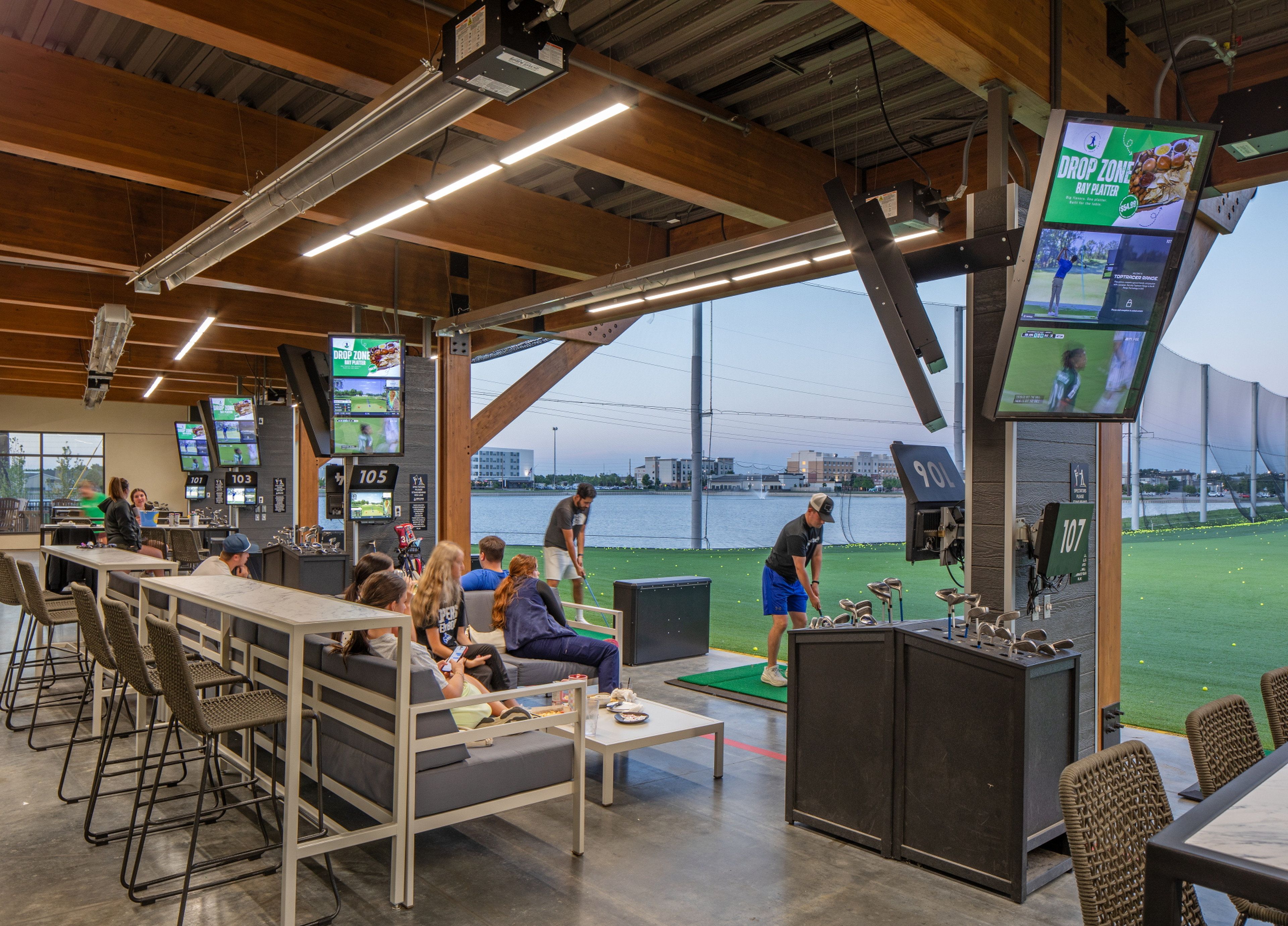 An indoor golf simulation area with multiple screens, seating, and a view of a golf course.
