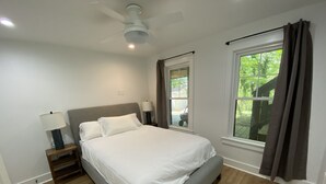 Un dormitorio con cama, mesitas de noche, ventilador de techo y ventanas con cortinas.