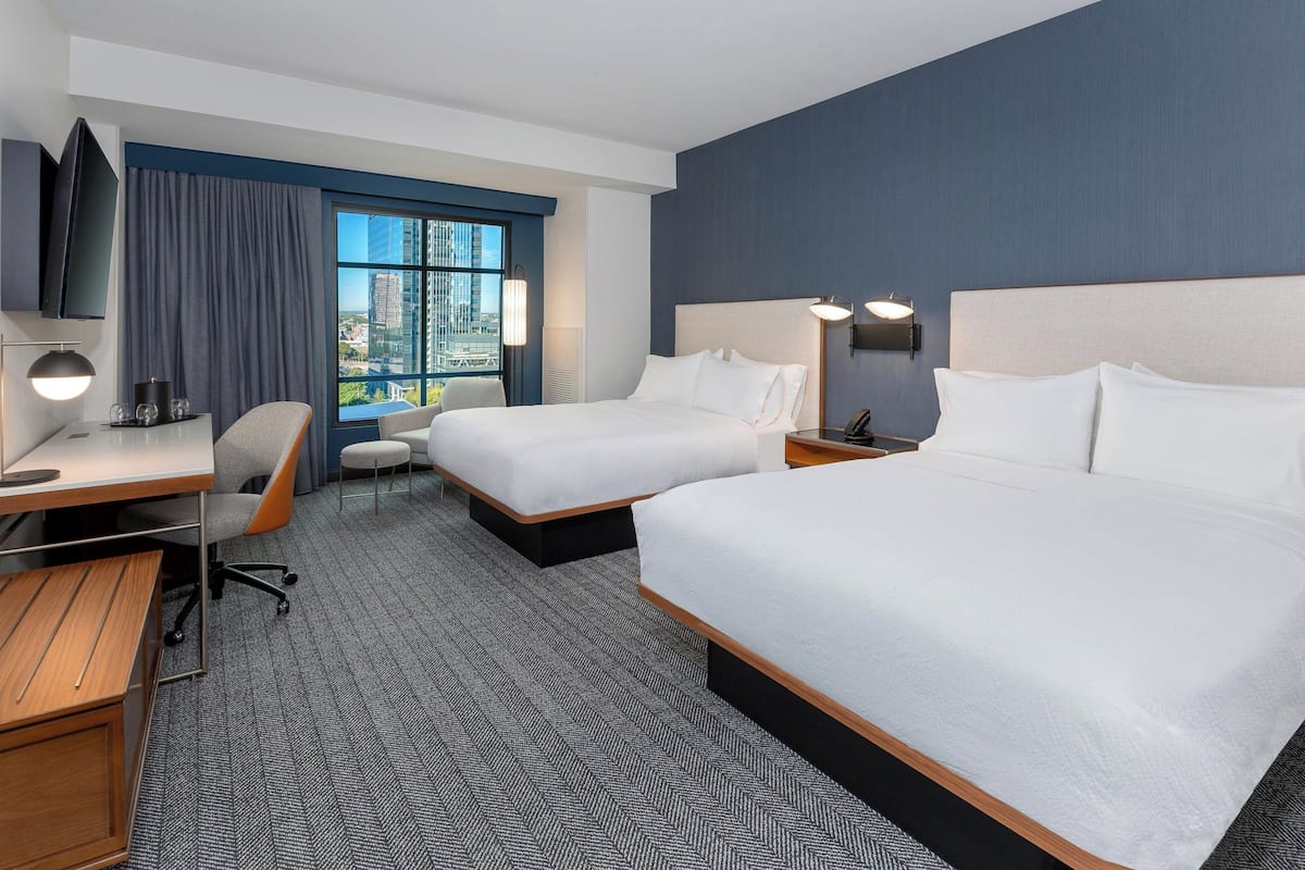 A hotel room with two beds, a desk, a chair, a TV, and a large window with a city view.