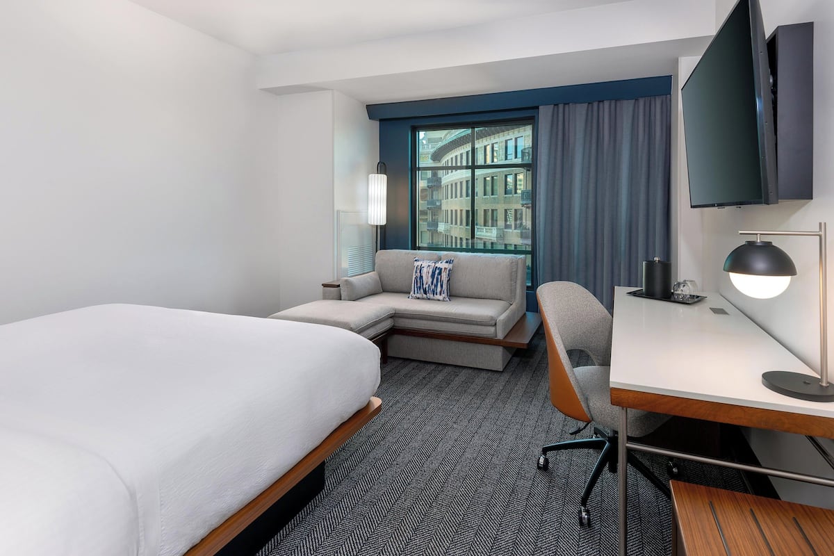 A hotel room with a bed, a sofa, a desk, a chair, a TV, and a window with a view of buildings.
