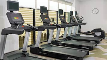 A row of treadmills with digital displays, arranged in a gym with large windows and a clock on the wall.