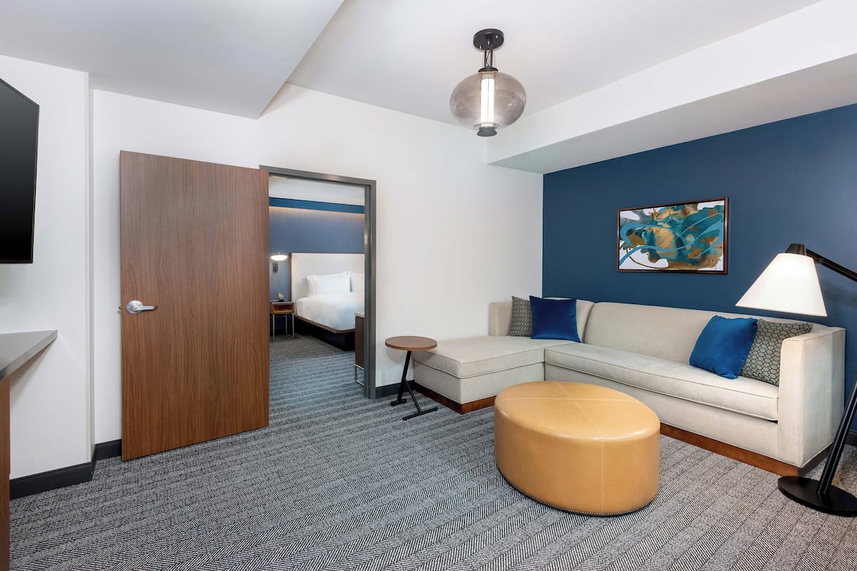 A modern hotel room with a wooden door, a sofa, a round ottoman, a lamp, and a painting on the wall.
