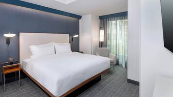 A hotel room with a large bed, a bedside table with a lamp, a chair, and a view of the outdoors through a window with curtains.