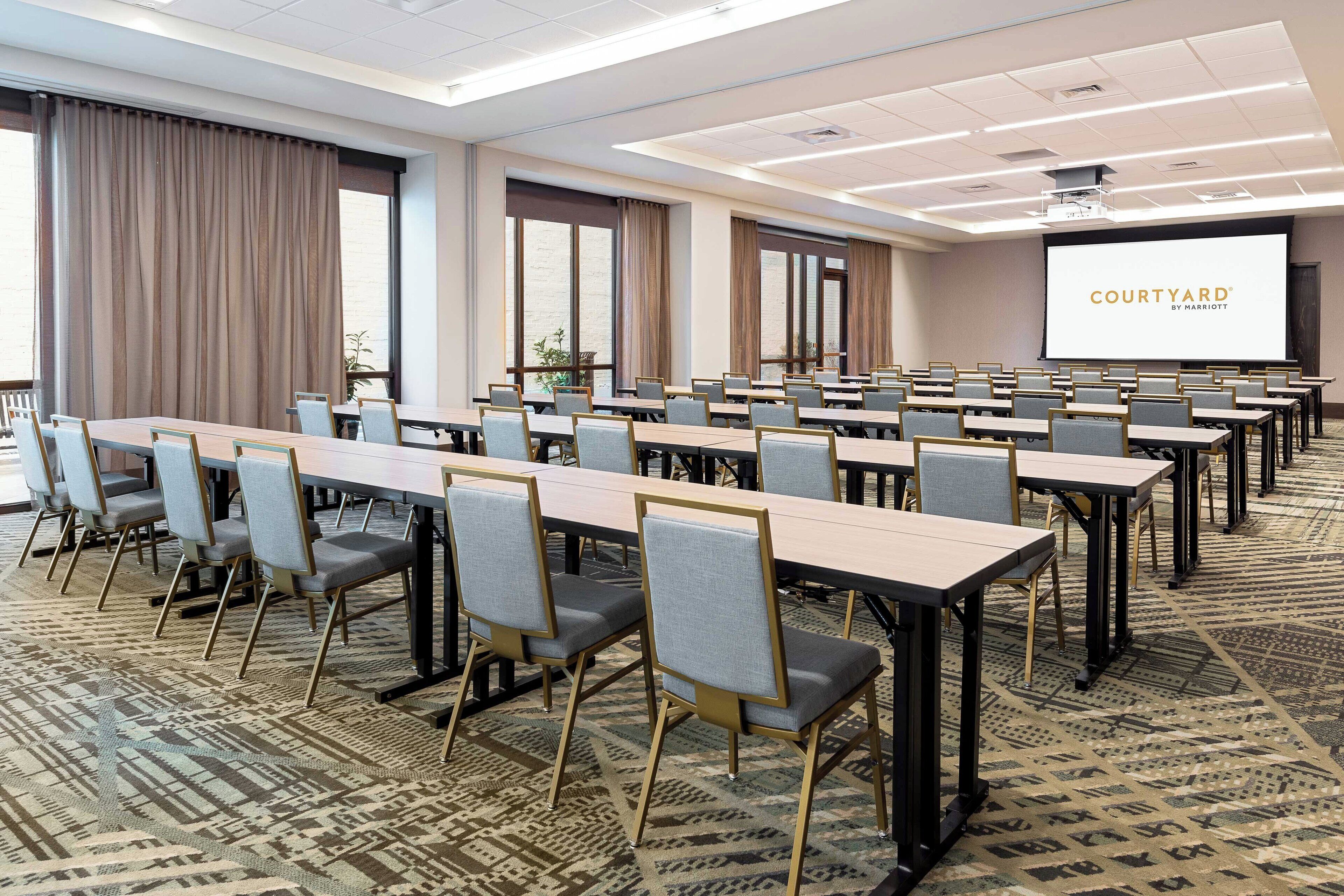 Uma sala de conferências com mesas e cadeiras compridas, uma tela grande exibindo 'COURTYARD BY MARHERTON' e carpete estampado.