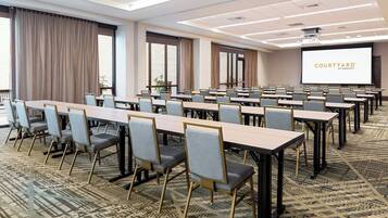Uma sala de conferências com mesas e cadeiras compridas, uma tela grande exibindo 'COURTYARD BY MARHERTON' e carpete estampado.