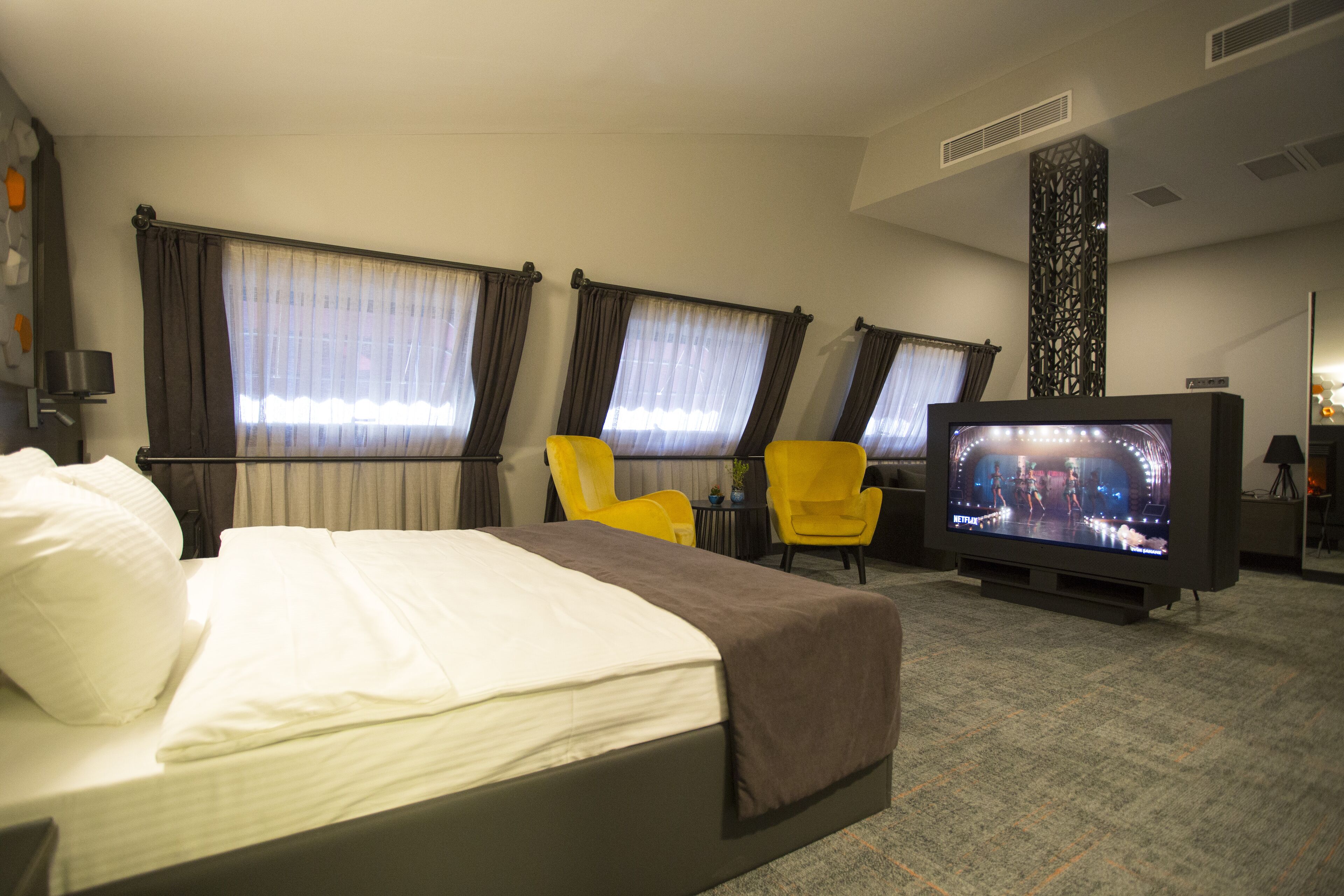 Une chambre d’hôtel avec un lit, deux chaises jaunes, une télévision et des rideaux aux fenêtres.
