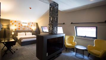 A hotel room with a bed, a flat-screen TV, a desk with a chair, two yellow armchairs, and a window with a view.