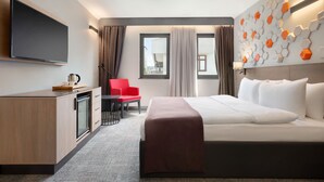 A hotel room with a bed, a desk with a flat-screen TV, a red chair, and a window with curtains.
