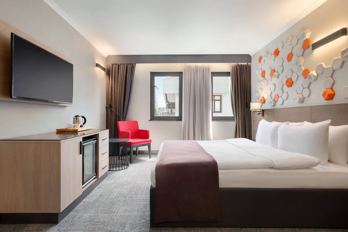Een hotelkamer met een bed, een bureau met een flatscreen televisie, een rode stoel en een raam met gordijnen.