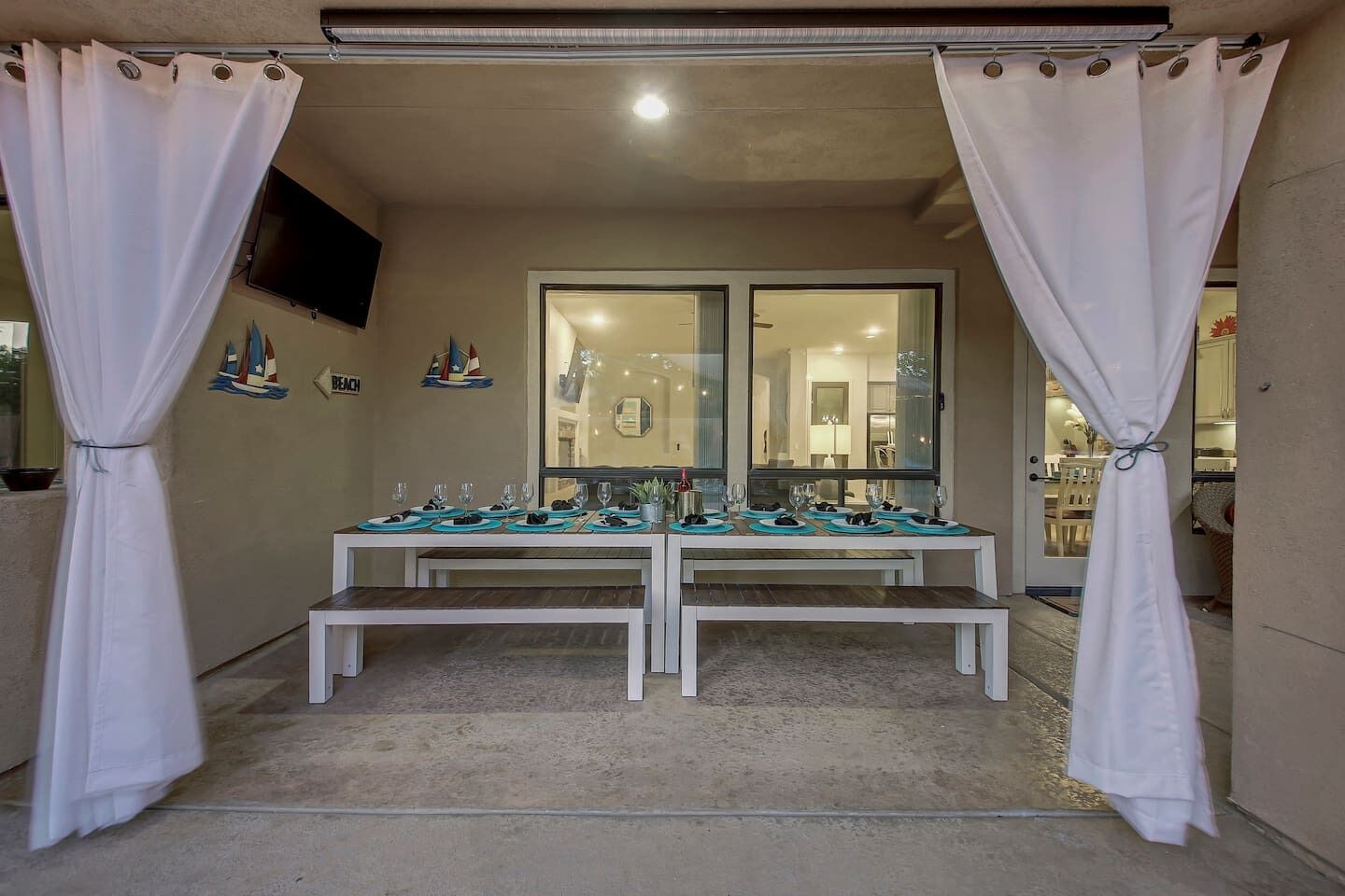 Un espace de restauration extérieur abrité, avec une table dressée pour quatre personnes, des rideaux blancs et une vue sur la pièce adjacente.