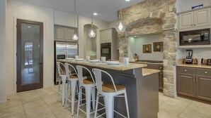A modern kitchen with a central island, bar stools, a stone accent wall, and built-in appliances. - Tahiti: Walking Distance to Coachella! (Indio)