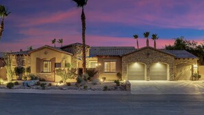 A two-story house with a stone facade, multiple garage doors, and a well-lit driveway. - Tahiti: Walking Distance to Coachella! (Indio)