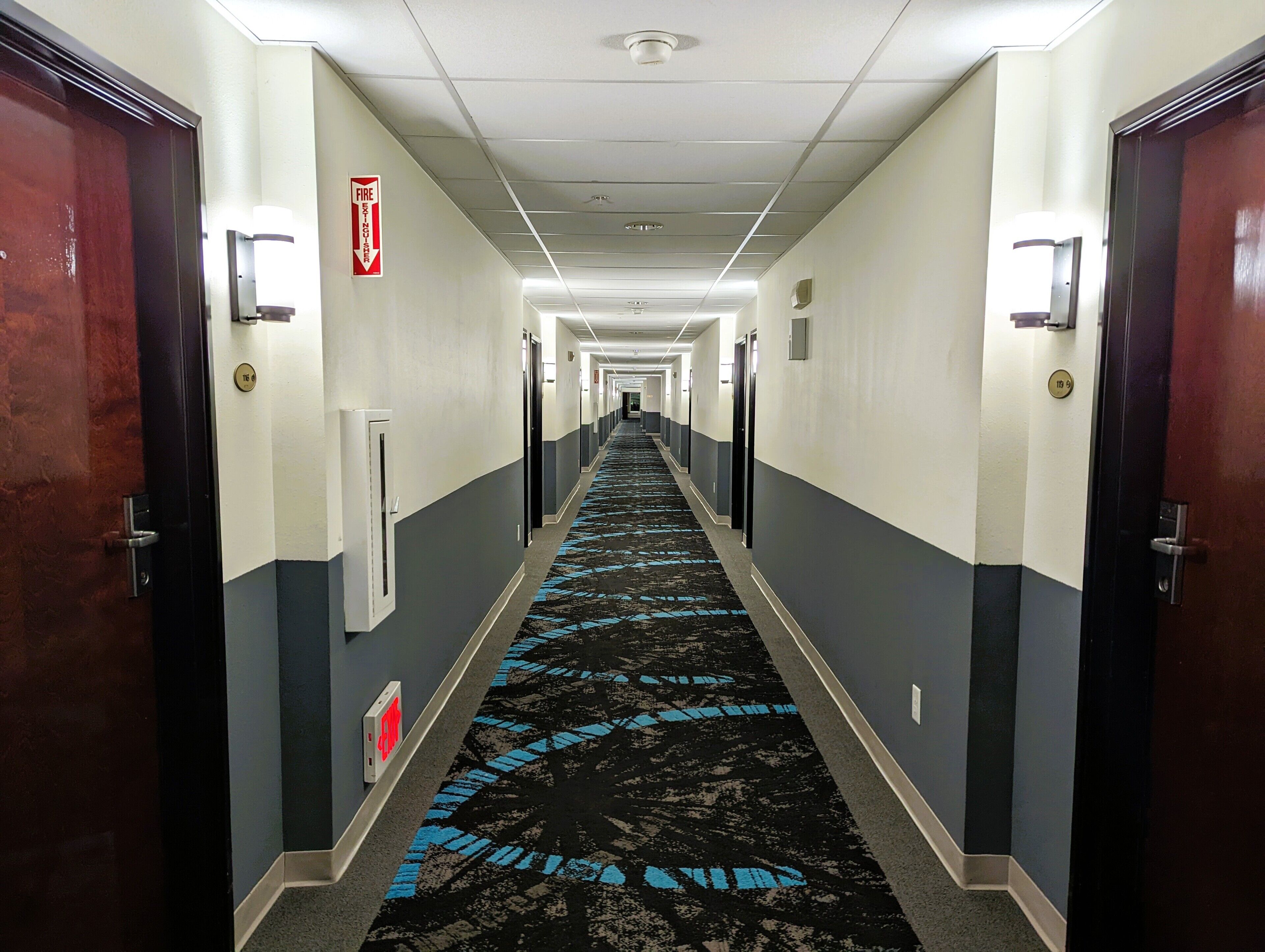 A hotel hallway with a blue carpet, doors on both sides, and a fire exit sign.