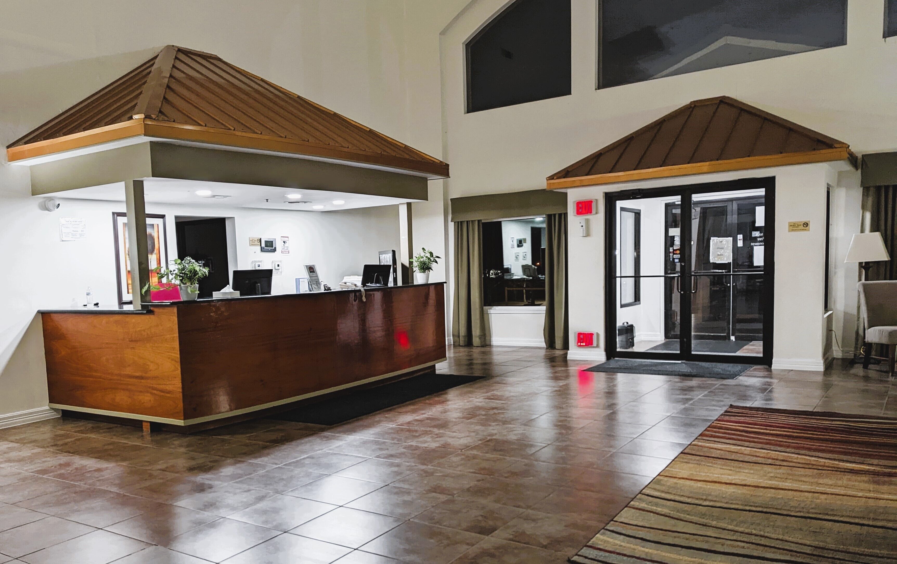 Un área de recepción con un mostrador de madera, un espacio de espera con un tapete estampado y una puerta de cristal de acceso.
