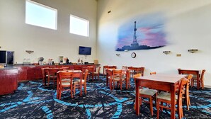 Uma área de refeições com mesas e cadeiras de madeira, uma cozinha com frigorífico e pia, e uma TV fixada na parede.