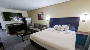 Quarto de hotel com uma cama grande, um sofá, uma mesa pequena, uma cozinha compacta com frigorífico e micro-ondas, e um quadro na parede.