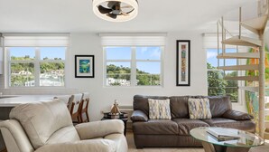 Ein modernes Wohnzimmer mit einem braunen Ledersofa, einem beige Armlehnstuhl und einem Glastisch. Zwei Fenster mit weißen Jalousien, ein Deckenventilator und gerahmte Kunstwerke an den Wänden sind zu sehen.