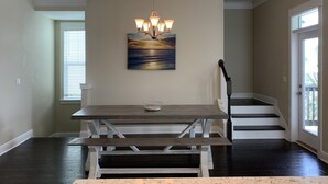Un coin repas avec une table et un banc en bois, un luminaire suspendu et un tableau représentant un coucher de soleil sur l’océan.