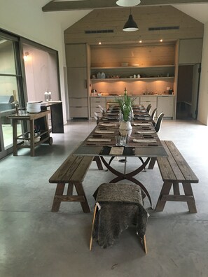 Un comedor amplio con una larga mesa de madera dispuesta para una comida, una mesita con un florero de follaje, y una zona de estar acogedora con una manta y un perro pequeño.