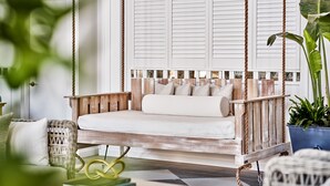 Un lit-balançoire en bois avec des coussins, une chaise en osier et une plante en pot, dans une pièce dotée d’une grande fenêtre et de volets.