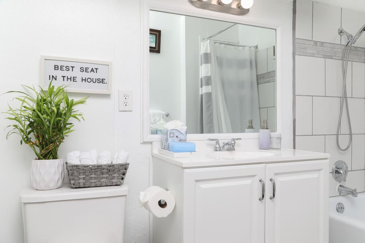 A bathroom with a white sink, a mirror, a shower, and a sign that reads "BEST SEAT IN THE HOUSE."