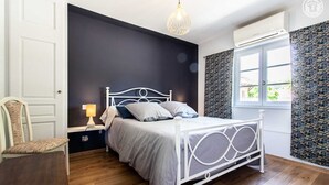 A bedroom with a white metal bed frame, a bedside table with a lamp, a chair, and a window with patterned curtains. - La Ferme de la Terve (BRUAILLES)