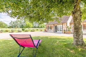 Une chaise pliante rouge est placée sous un arbre, dans un espace verdoyant, avec une maison en arrière-plan.