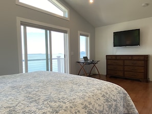Un dormitorio con una cama grande, un escritorio con una computadora portátil, un televisor en la pared y vista al océano a través de las ventanas.