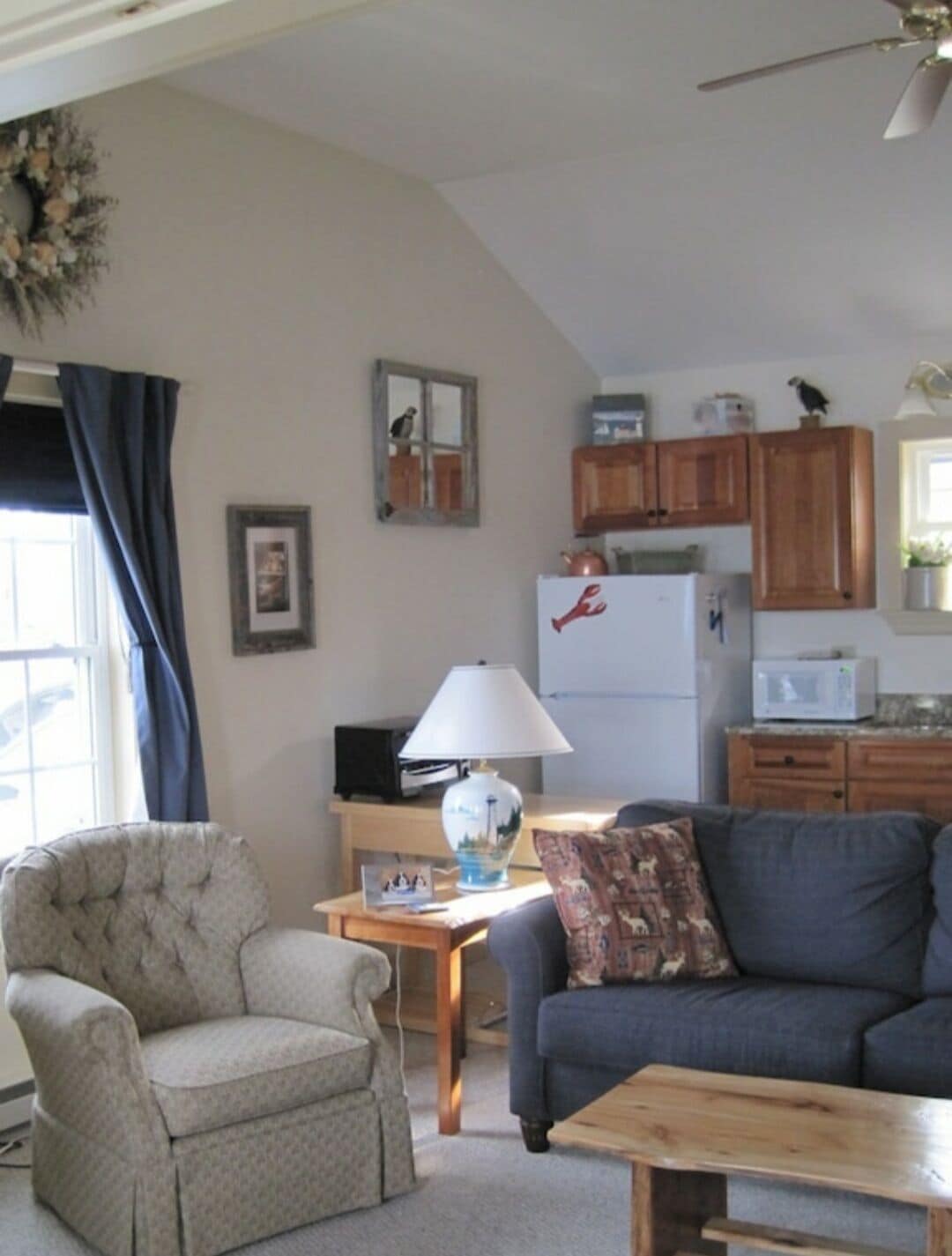 Un salon chaleureux avec un fauteuil gris, un canapé et une table basse en bois. Une lampe et un cadre photo sont posés sur une table d’appoint. La cuisine, visible à l’arrière-plan, est équipée de meubles en bois et d’un four à micro-ondes.