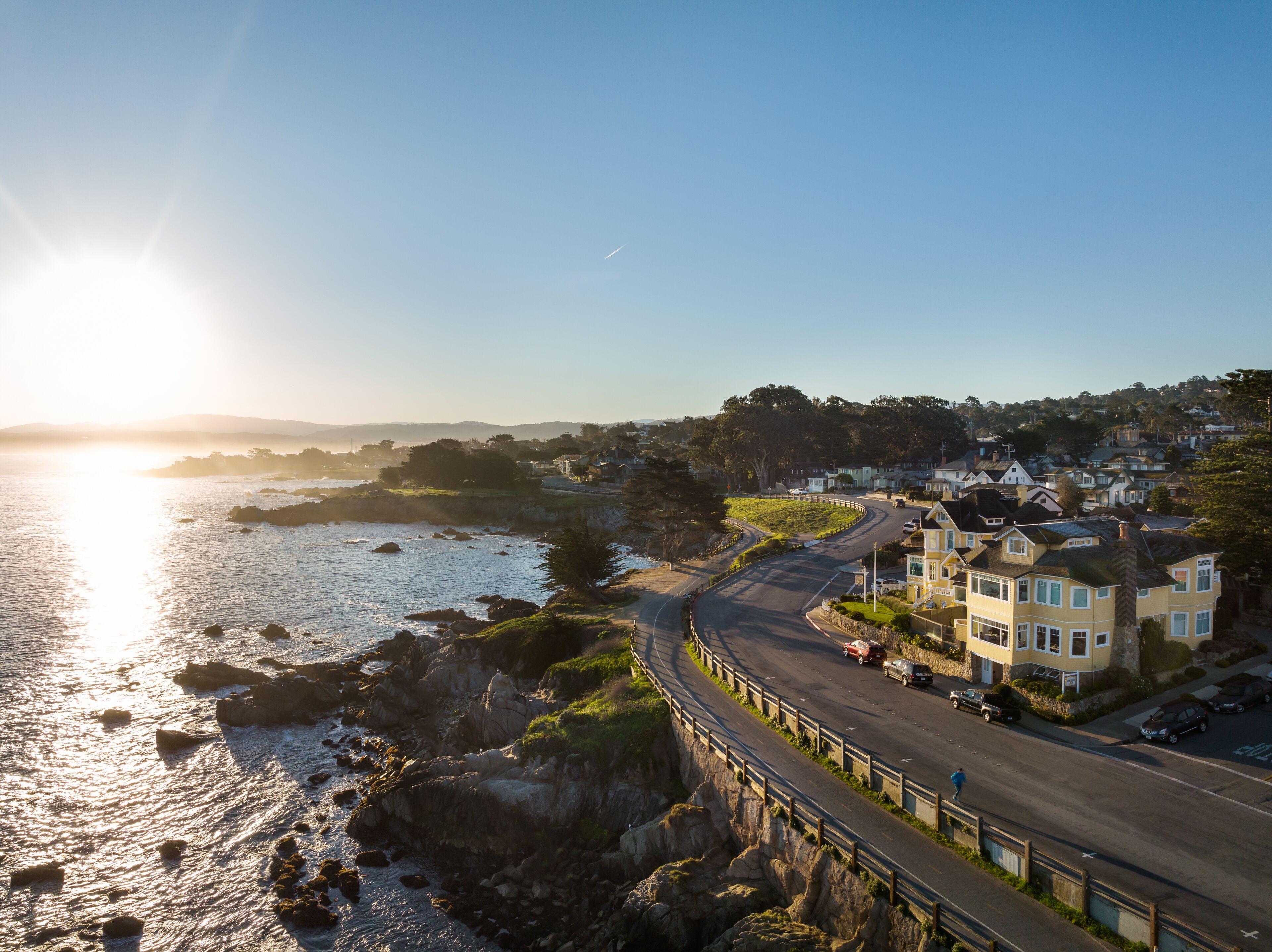 A coastal road curving along a rocky shoreline with houses on one side and a clear view of the ocean.