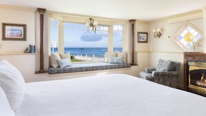 A bedroom with a large bed, a window seat with a view of the ocean, a fireplace, and a chair. - Seven Gables Inn on Monterey Bay, A Kirkwood Collection Property (Pacific Grove)