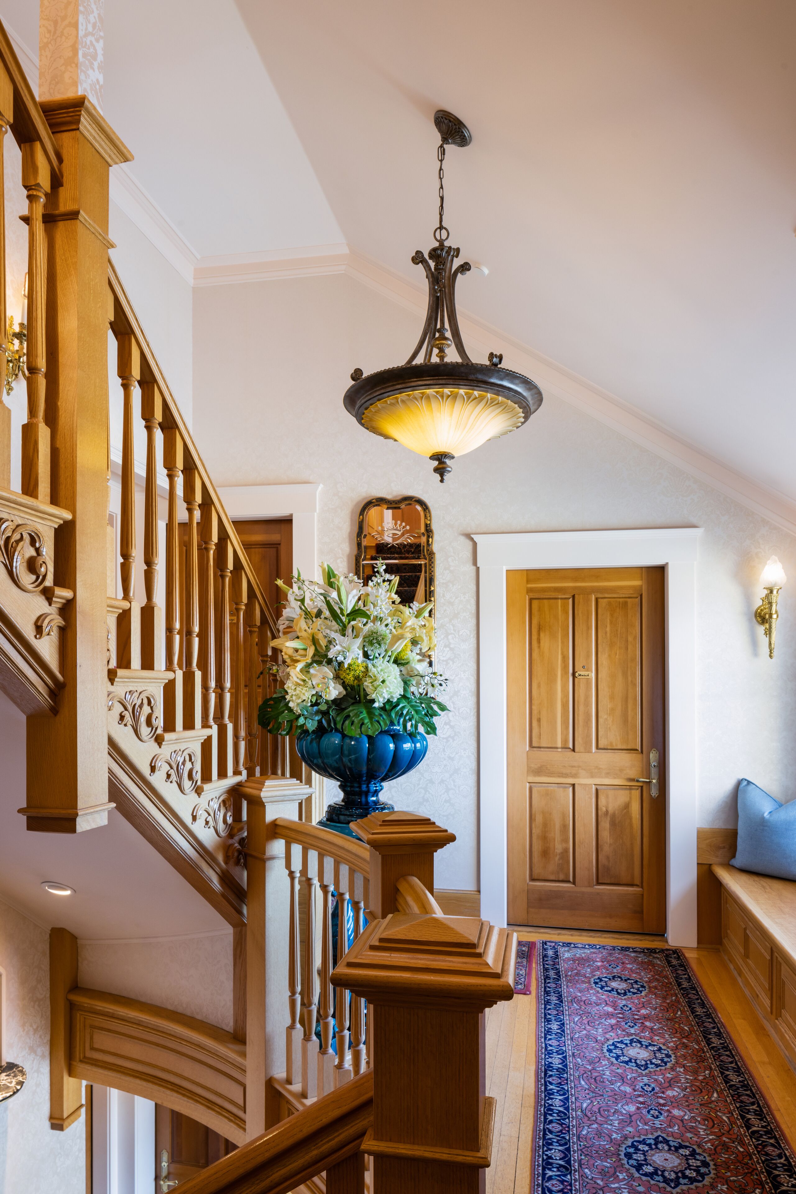 A wooden staircase with a blue vase of flowers, a patterned rug, and a wooden door at the top.