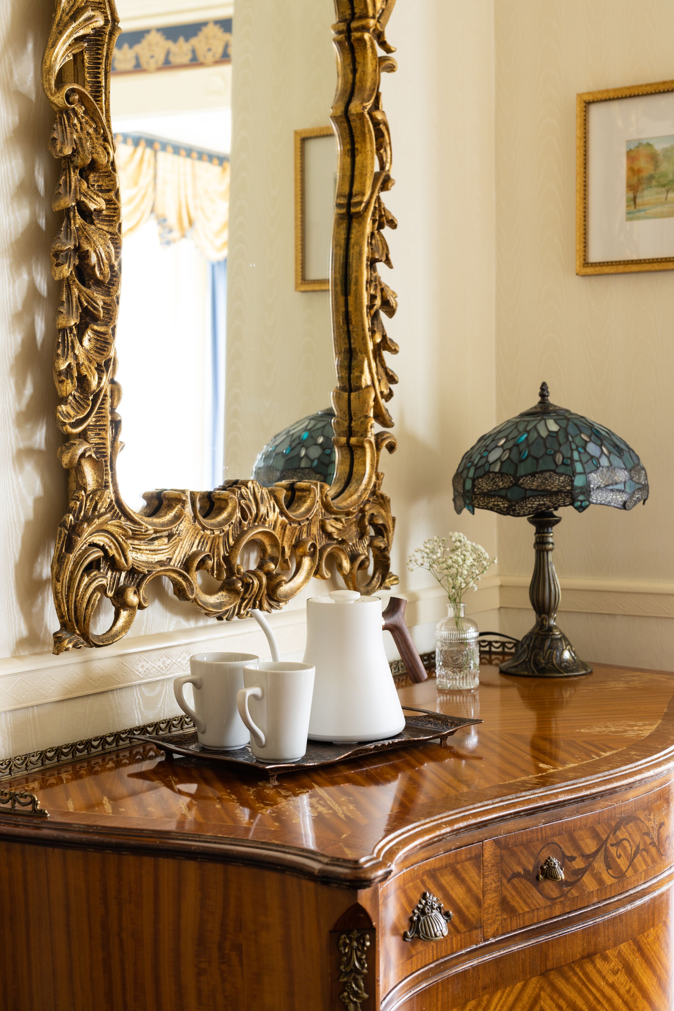 A wooden dresser with a gold ornate mirror, a white pitcher, and a green stained glass lamp.