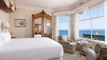 A hotel room with a large bed, a sitting area with patterned chairs, a round table, a mirror, and a view of the sea through large windows.