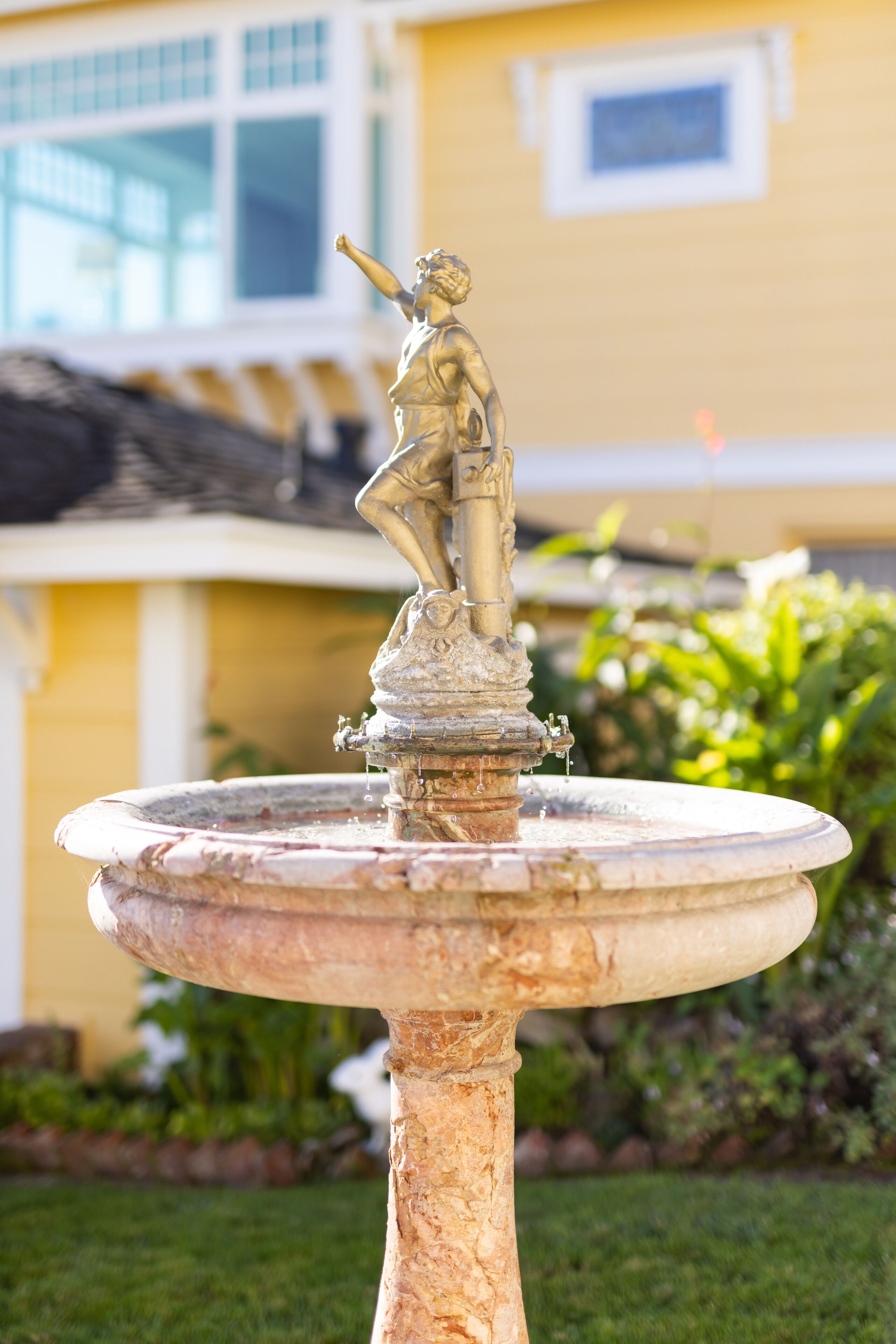 A bronze statue on a fountain in a garden with a yellow building in the background.