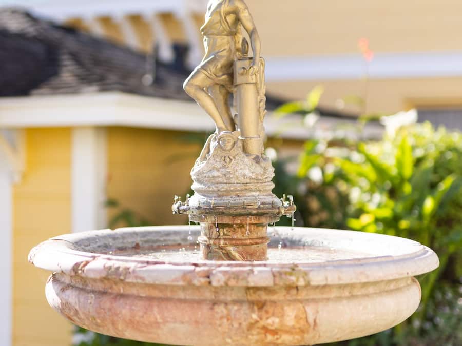 A bronze statue on a fountain in a garden with a yellow building in the background.