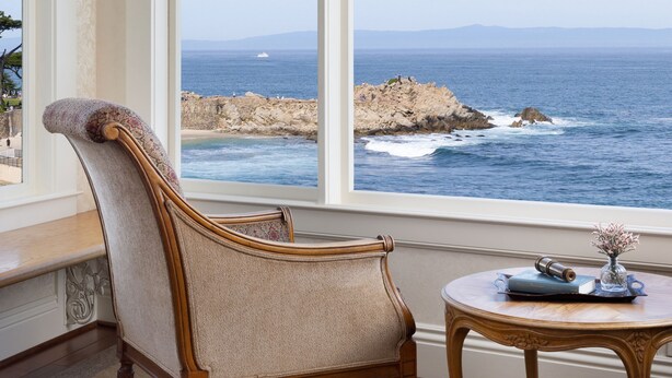 A room with a large window overlooking the sea, a wooden chair, a small round table, and a chandelier. - Seven Gables Inn on Monterey Bay, A Kirkwood Collection Property (Pacific Grove)