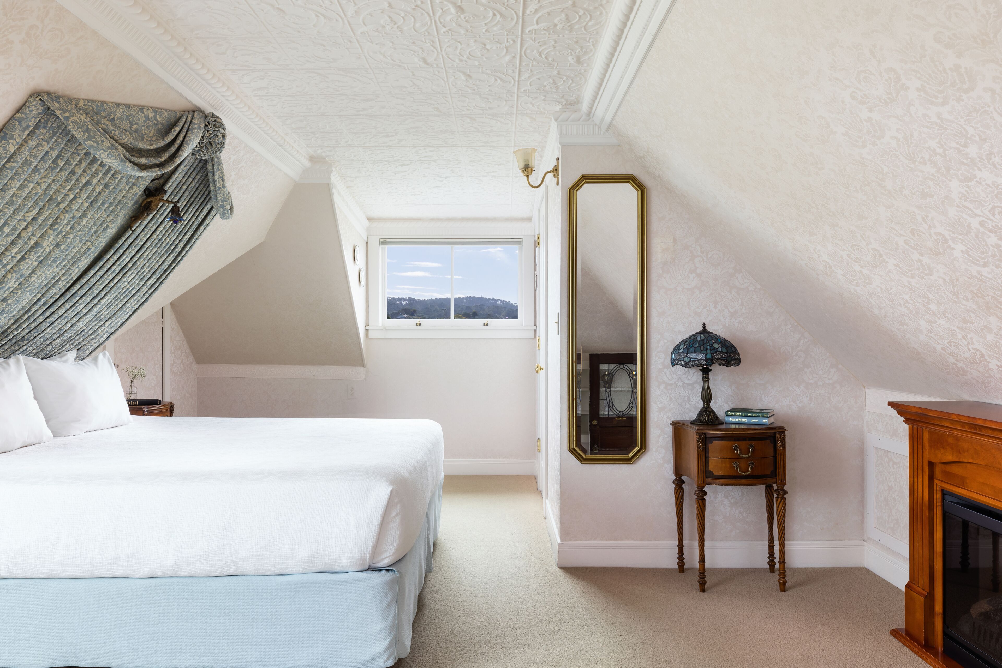 A bedroom with a large bed, a mirror, a small table with a lamp, and a fireplace.