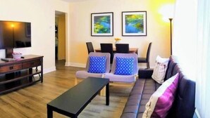 Un salon moderne avec un téléviseur à écran plat, une table basse en bois et un canapé agrémenté de coussins à motifs. Deux œuvres d’art encadrées ornent le mur, et un luminaire est fixé au plafond.