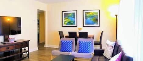 A modern living room with a flat-screen TV, a wooden coffee table, and a sofa with patterned cushions. There are two framed artworks on the wall and a ceiling light fixture.
