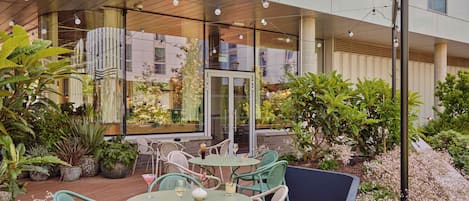 Un patio exterior con mesas y sillas redondas, rodeado de macetas con plantas y arbustos florecientes.