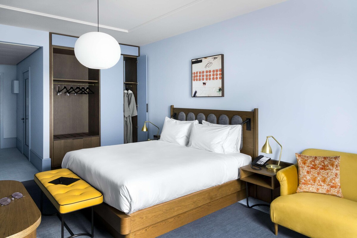 A hotel room with a wooden bed, a yellow bench, a yellow armchair, a bedside table with a lamp, and a coat rack with clothes.