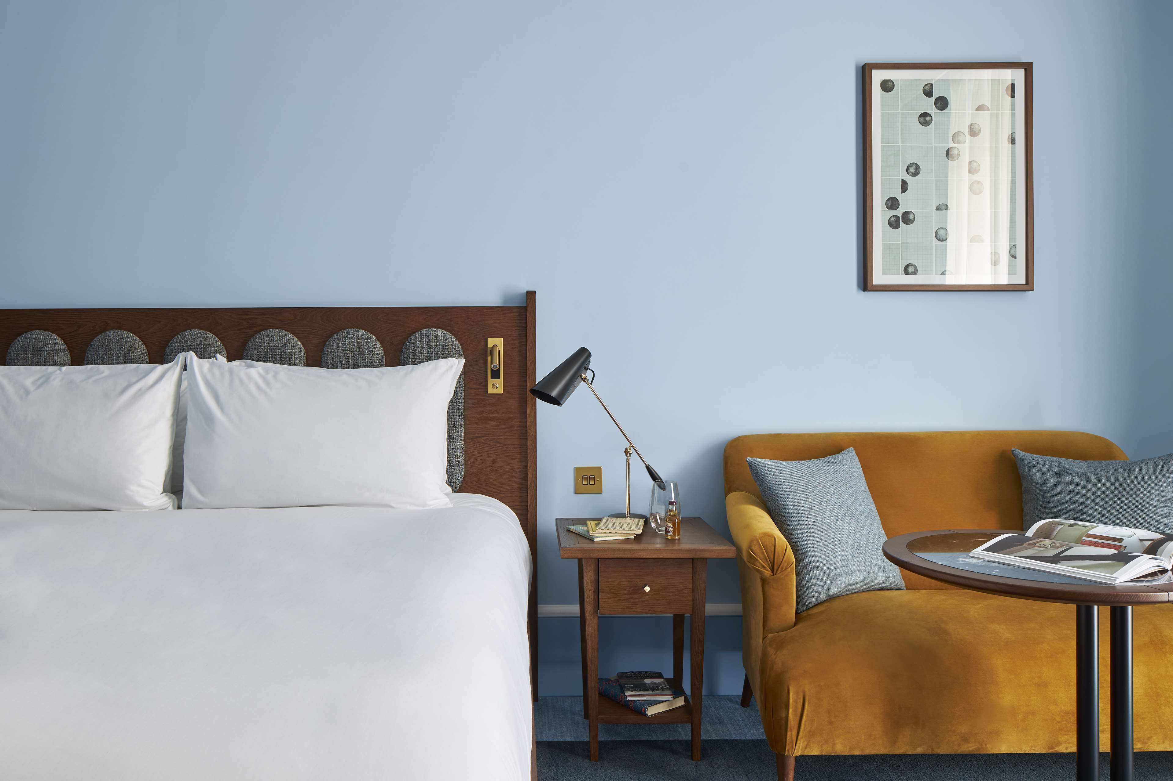 A hotel room with a bed, a nightstand with a lamp, a small table with a magazine, and a sofa.