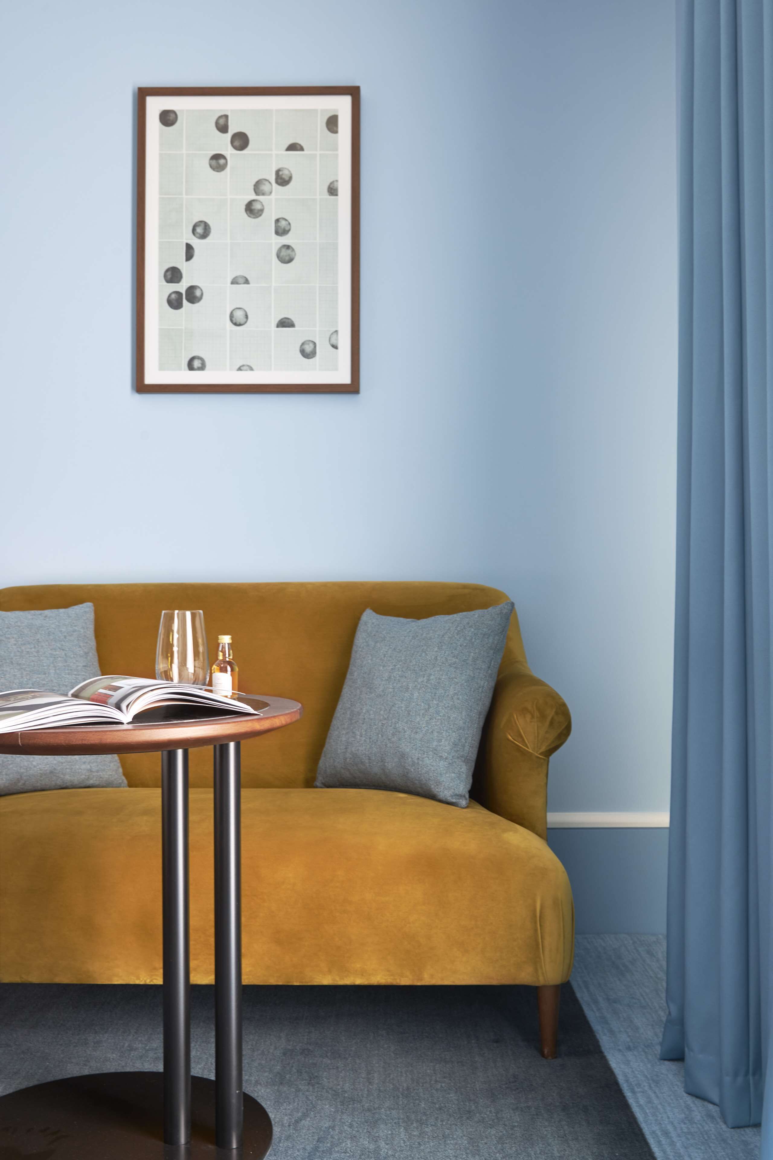 A room with a mustard-colored sofa, a round table with a book and a glass, and a framed artwork on the wall.
