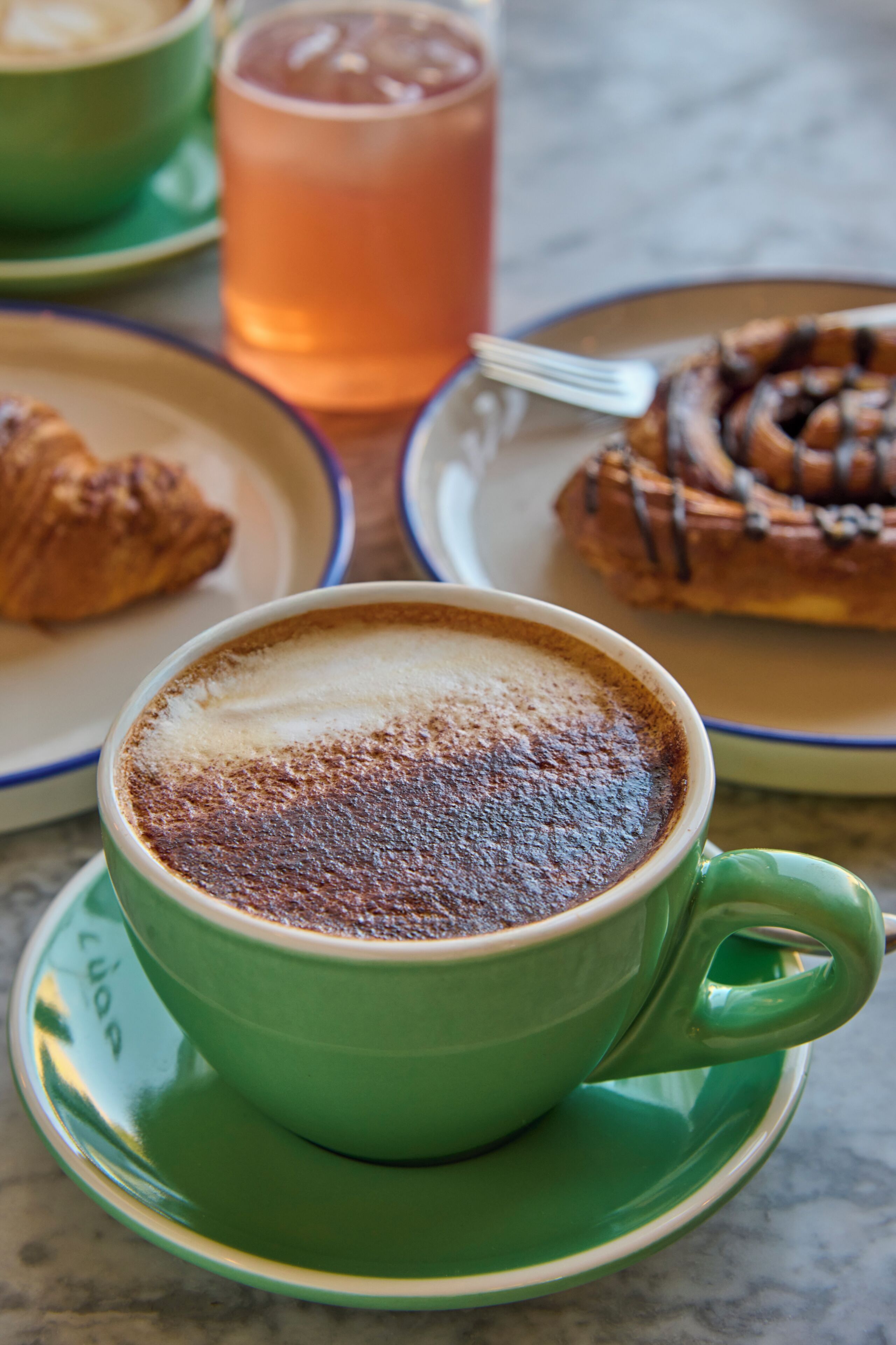 A cup of coffee with a frothy top, a pastry, and a glass of iced tea on a table.